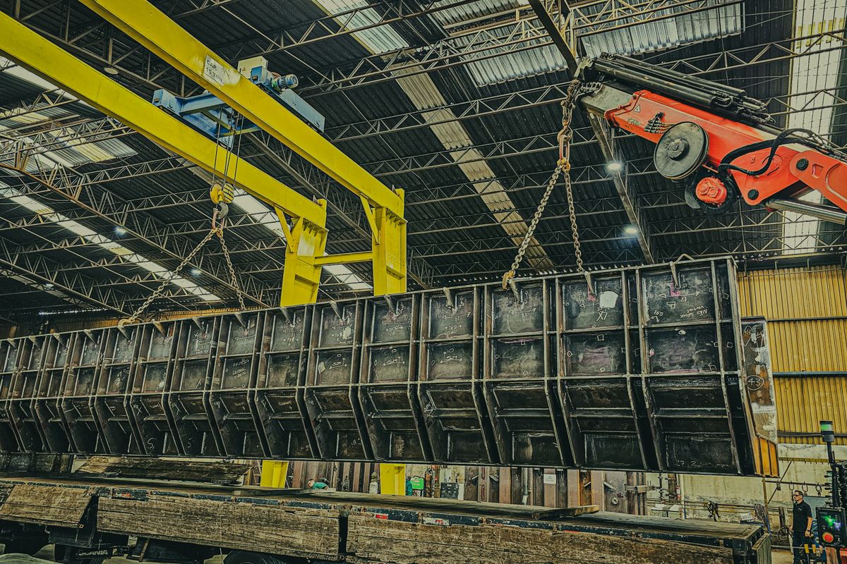 Heavy steel structure being lifted by cranes during warehouse construction and component fabrication.